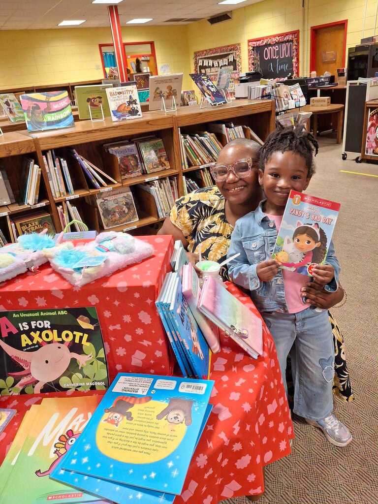 A woman and a young girl sit at a table with a variety of books in a library.