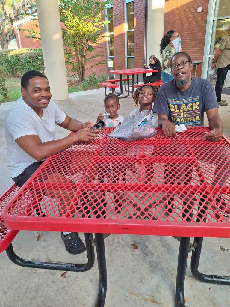 Three people sit at a red picnic table, one holding a child and the other two smiling. A building is in the background.