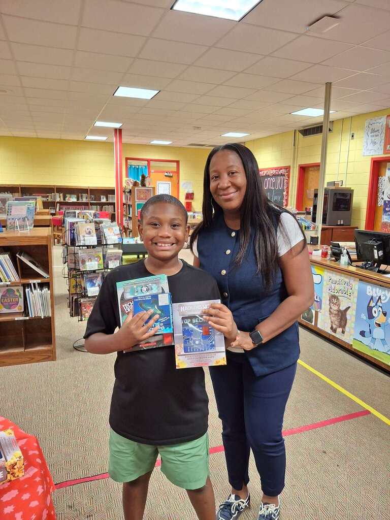 A boy and an adult stand in a library, holding books, with shelves, tables, and a yellow floor line.