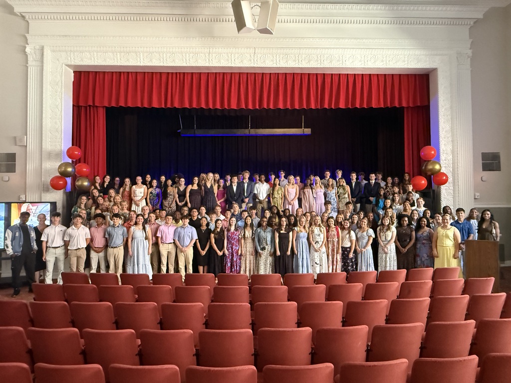 A group of students stands on a stage with a red curtain, red chairs in rows, and balloons.