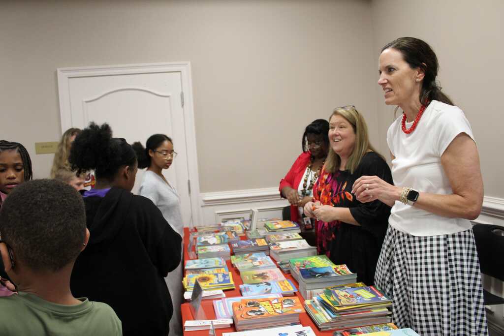 A woman stands by a table with books, surrounded by children and adults. They look at the books.
