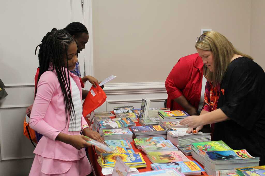 A woman in black and another in red look at books. A girl in pink holds a book. A table has multiple books.