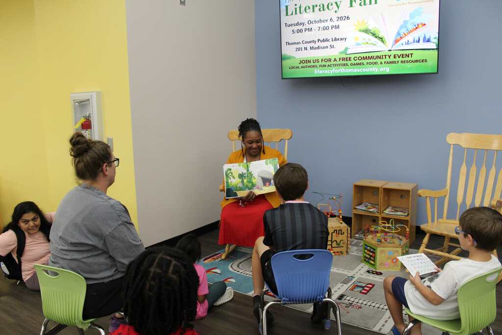 Woman reads a book to children in chairs in a room with a green screen and a sign.