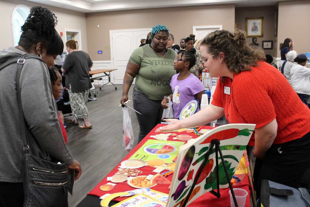 A group of people inside a room with a table full of colorful items, one person showing something to others.
