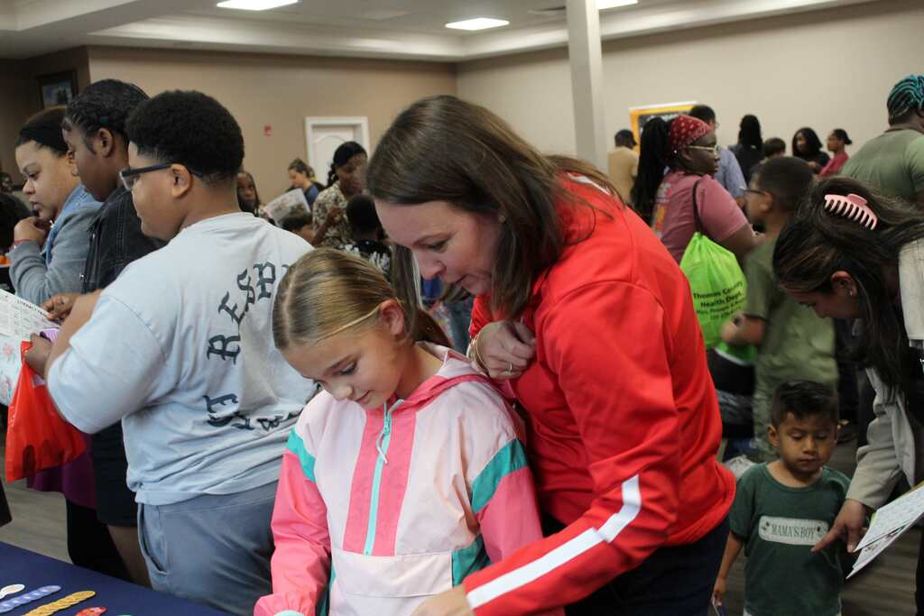 A woman helps a child in a red jacket and pink and white jacket. Behind them, many people stand and interact in a room.
