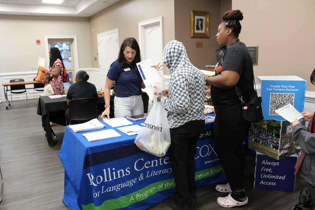 Three women stand by a table with a blue cover, reading material. Behind them, others sit and stand.