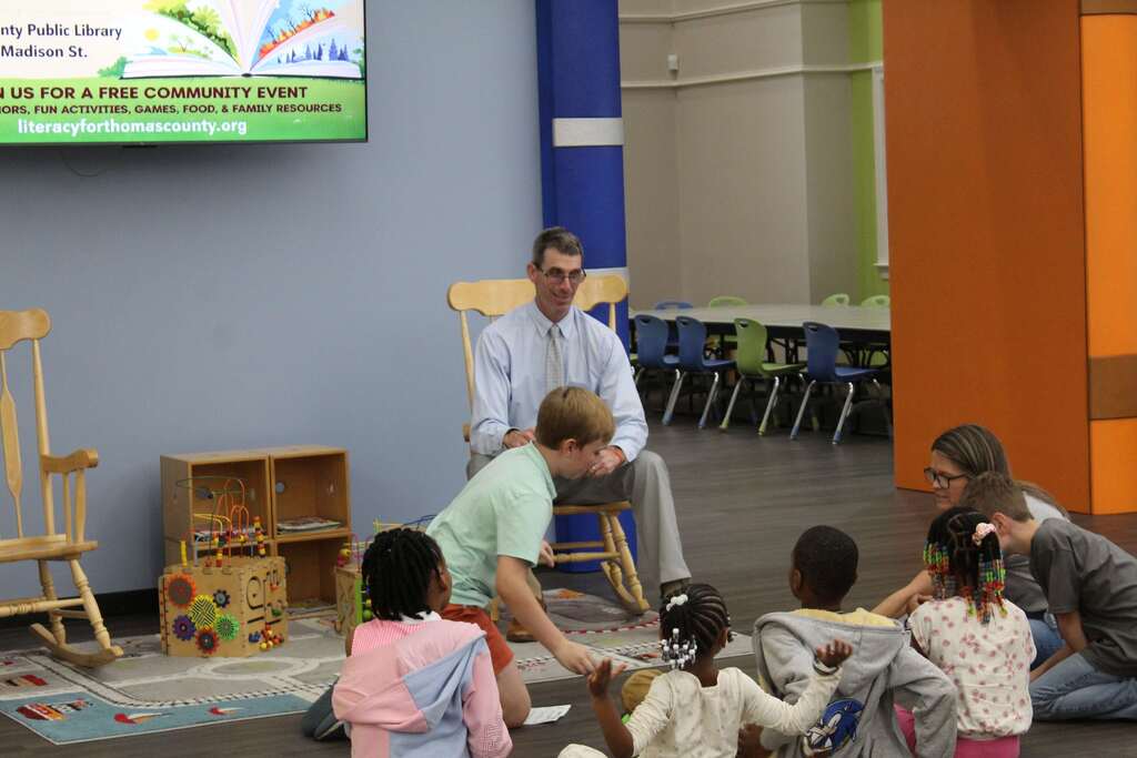 A man in a suit leads a story time for children sitting on the floor, chairs around a small stage.