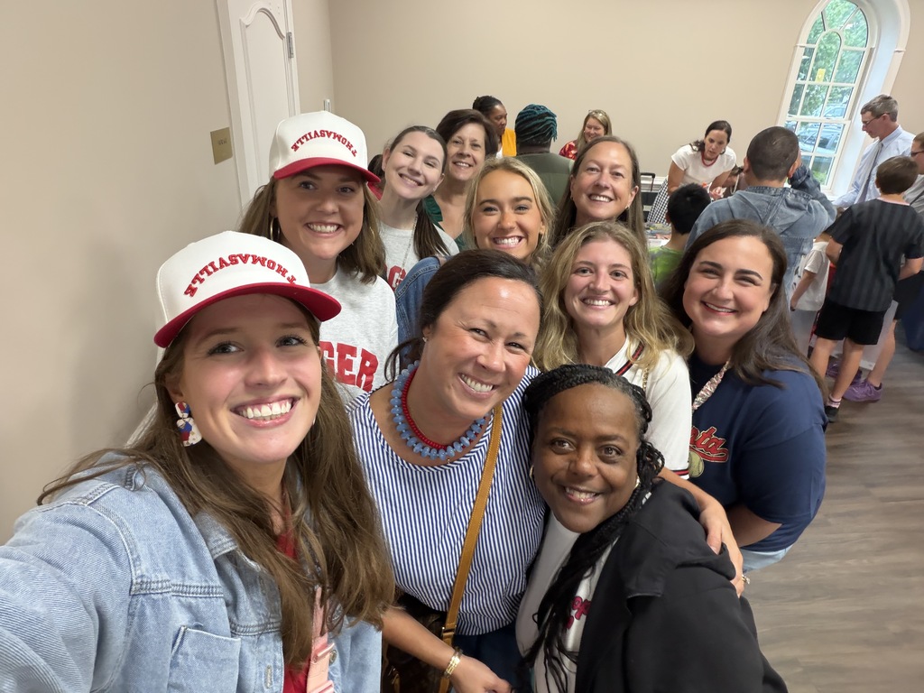 Group of smiling women and girls wearing hats with "St. Thomas" written on them. They are standing in a room with wood flooring.