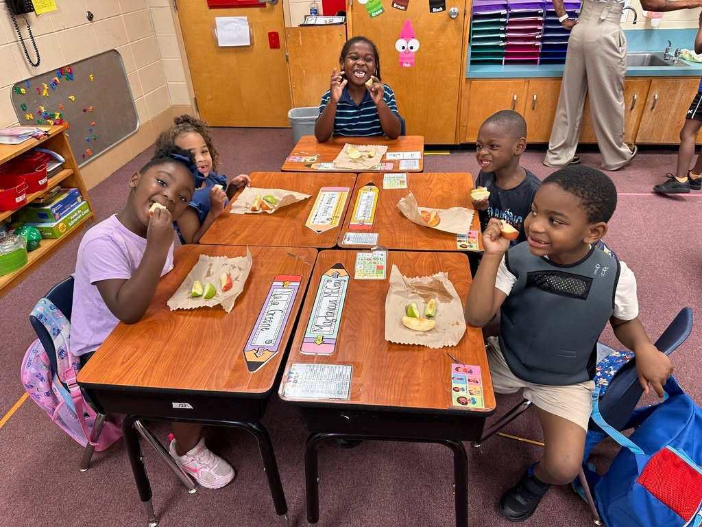 A group of children seated at desks in a classroom, eating sandwiches. A person stands in the background.