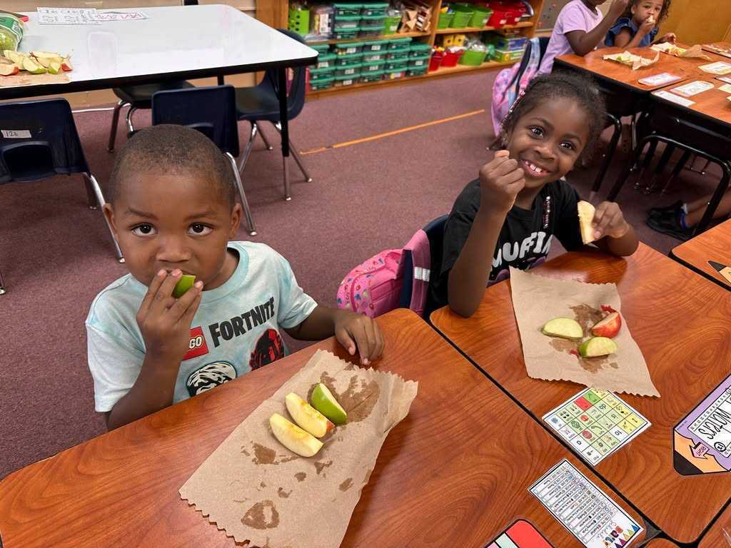 Two young children eat fruit slices at desks. A boy holds a piece in his hand, and a girl smiles.
