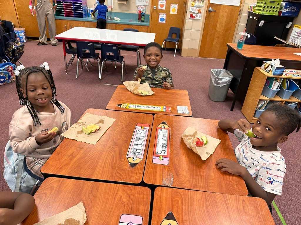 Three children sit at desks, eating sandwiches. Behind them are more desks and chairs. The room has wooden floors and walls.