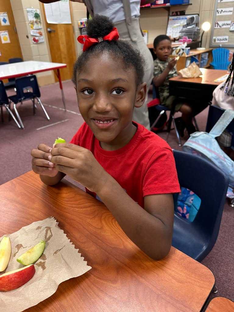 A child in a red shirt sits at a table eating an apple slice. A blurred background shows other children sitting at desks.