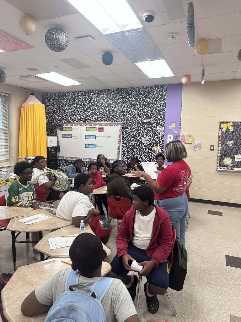 A classroom with students seated at desks. An adult stands at the front, addressing them. Walls display educational materials.