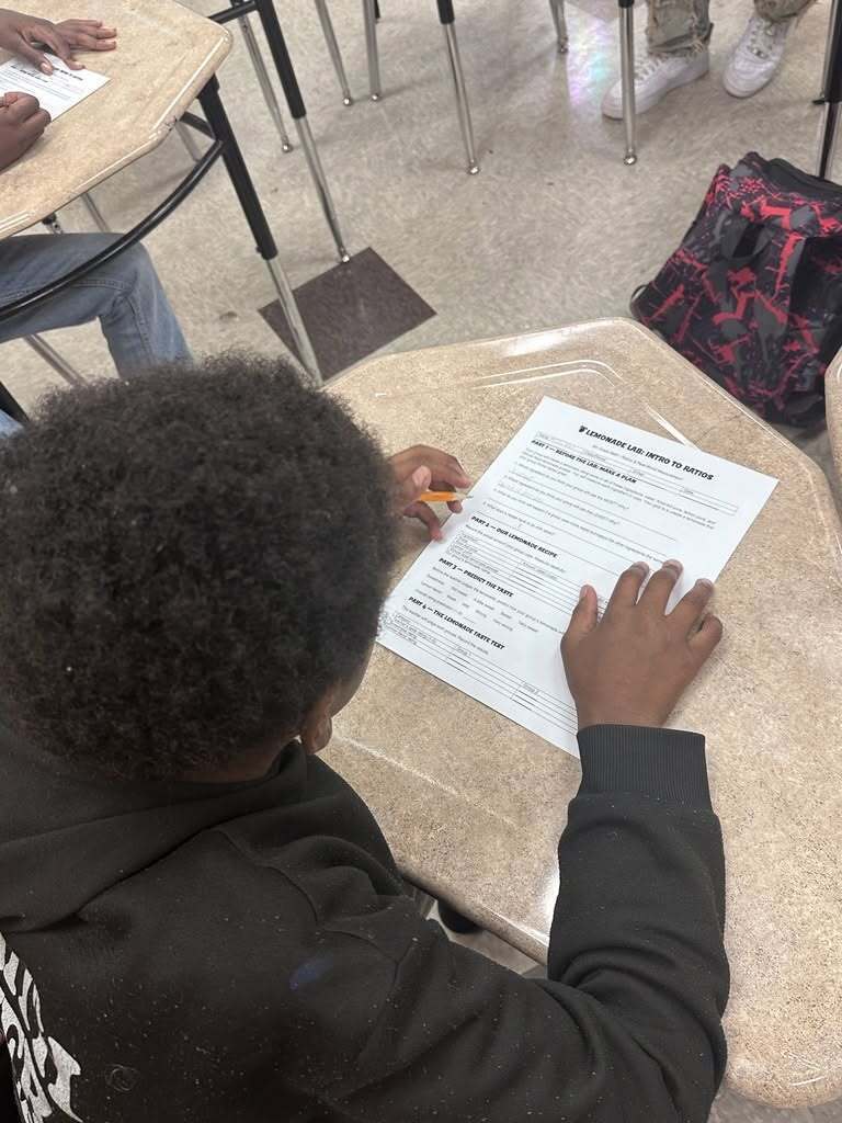 A student fills out a form at a desk. Other students sit in the background.