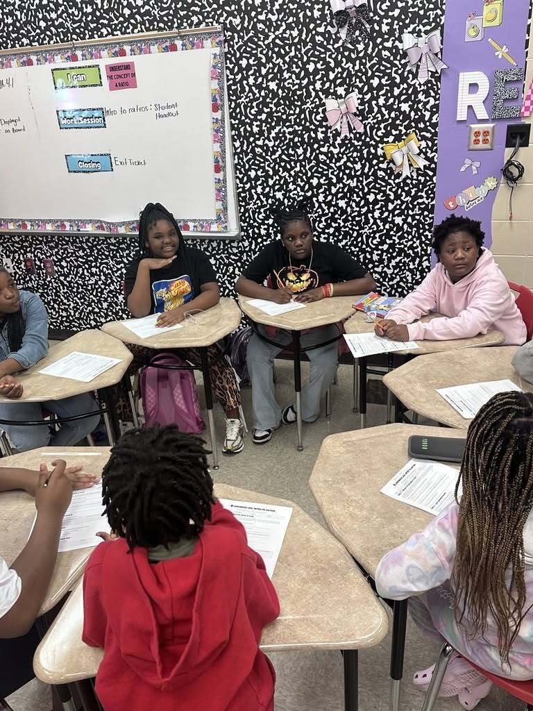 Students seated around desks in a classroom, facing forward and writing. A whiteboard and decorations are in the background.