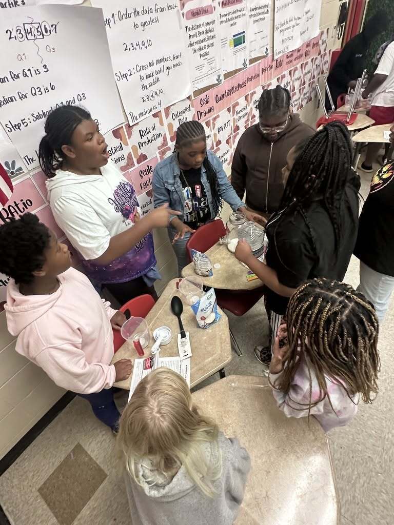 Several young people gather around tables in a classroom, engaged in an activity with spoons, bowls, and paper.