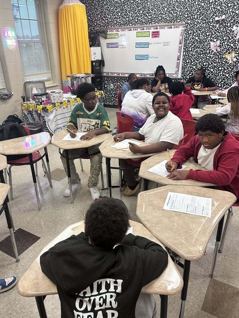 Students seated at desks in a classroom. They have writing materials and papers in front of them.