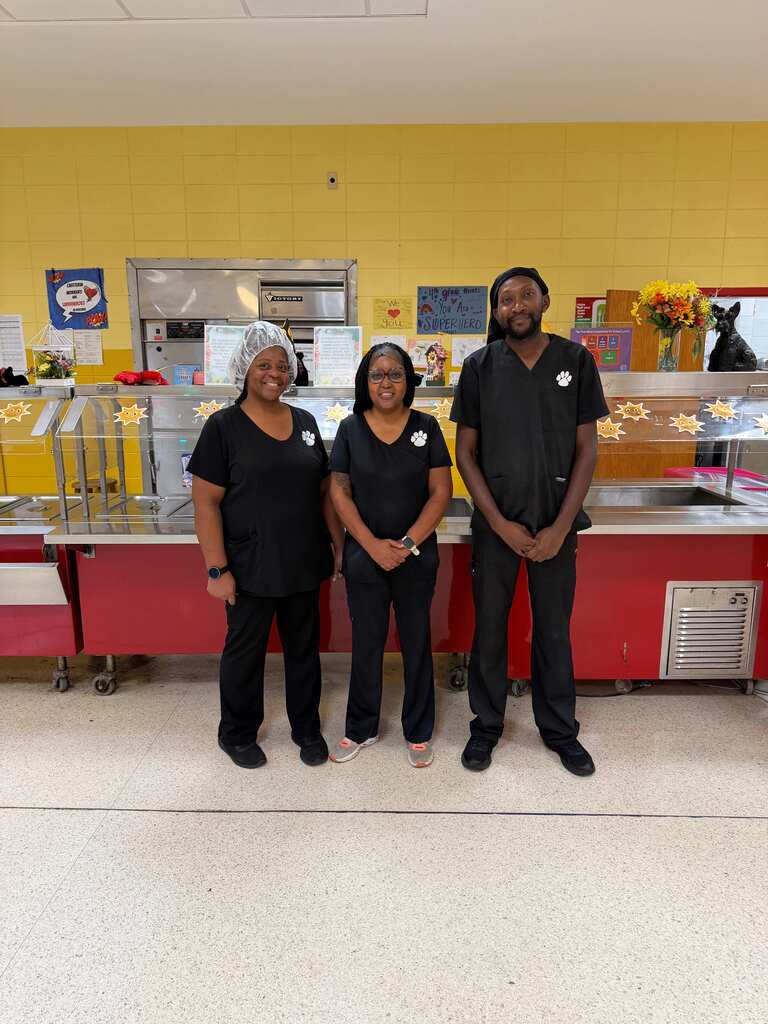 Three people in black uniforms pose together in a room with yellow walls and a counter.
