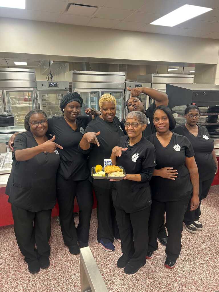 Workers in matching uniforms posing for a photo in a kitchen with trays of food.