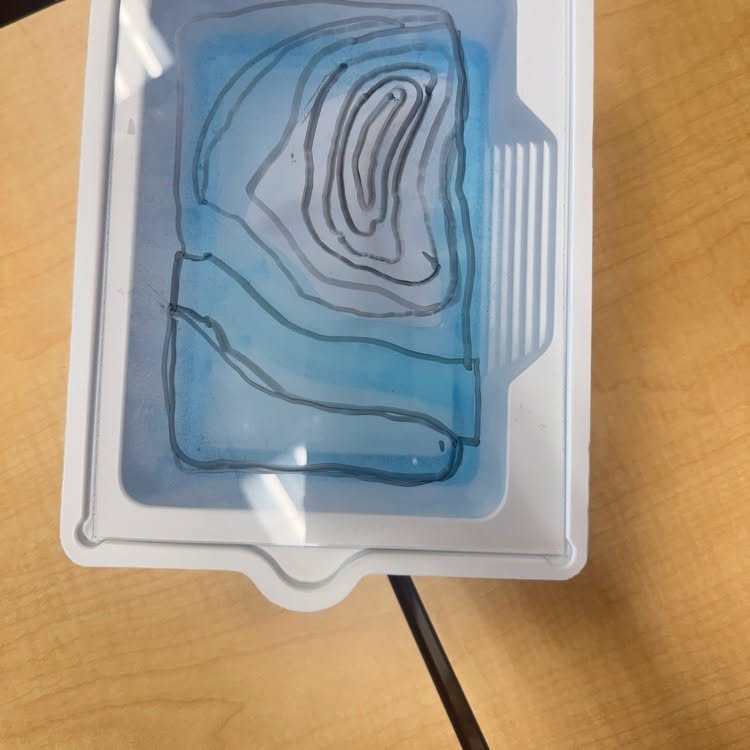 A clear plastic container on a wooden surface holds a blue liquid with a line pattern resembling a map.