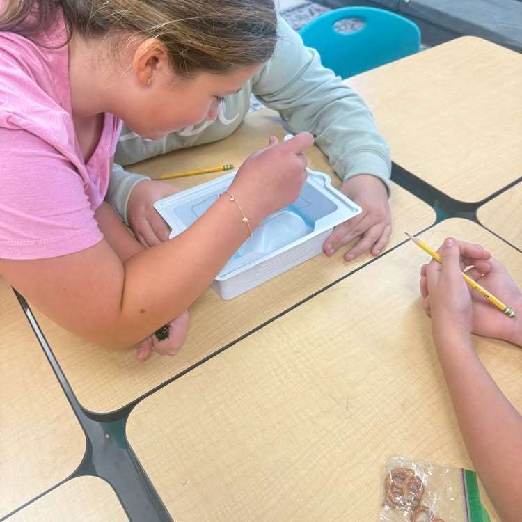 A person in pink works on a project in a box with a pencil. Another person helps.