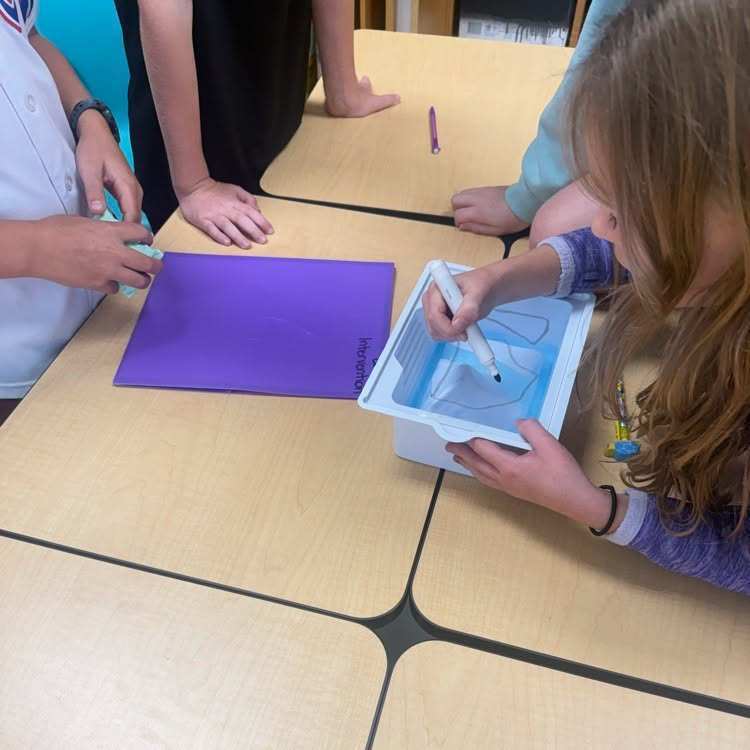 A group of children in a classroom, one of them drawing on a plastic board.