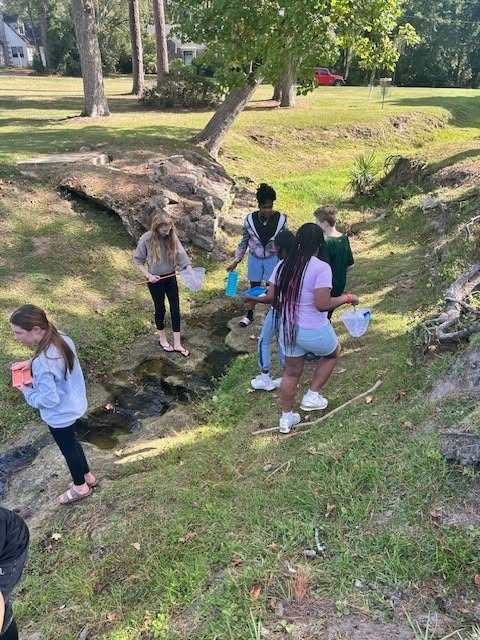 Five people are outside near a stream with trees in the background. They hold cups and examine water.
