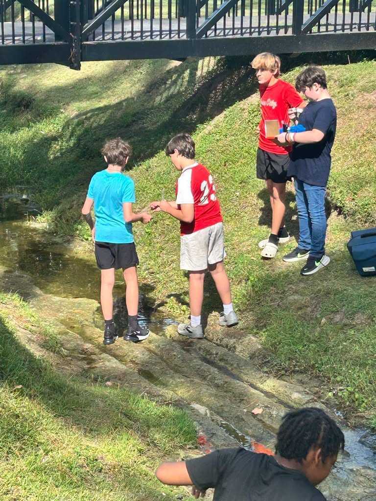 Six boys outside near a water source. One holding a clear container. They are standing on grass.