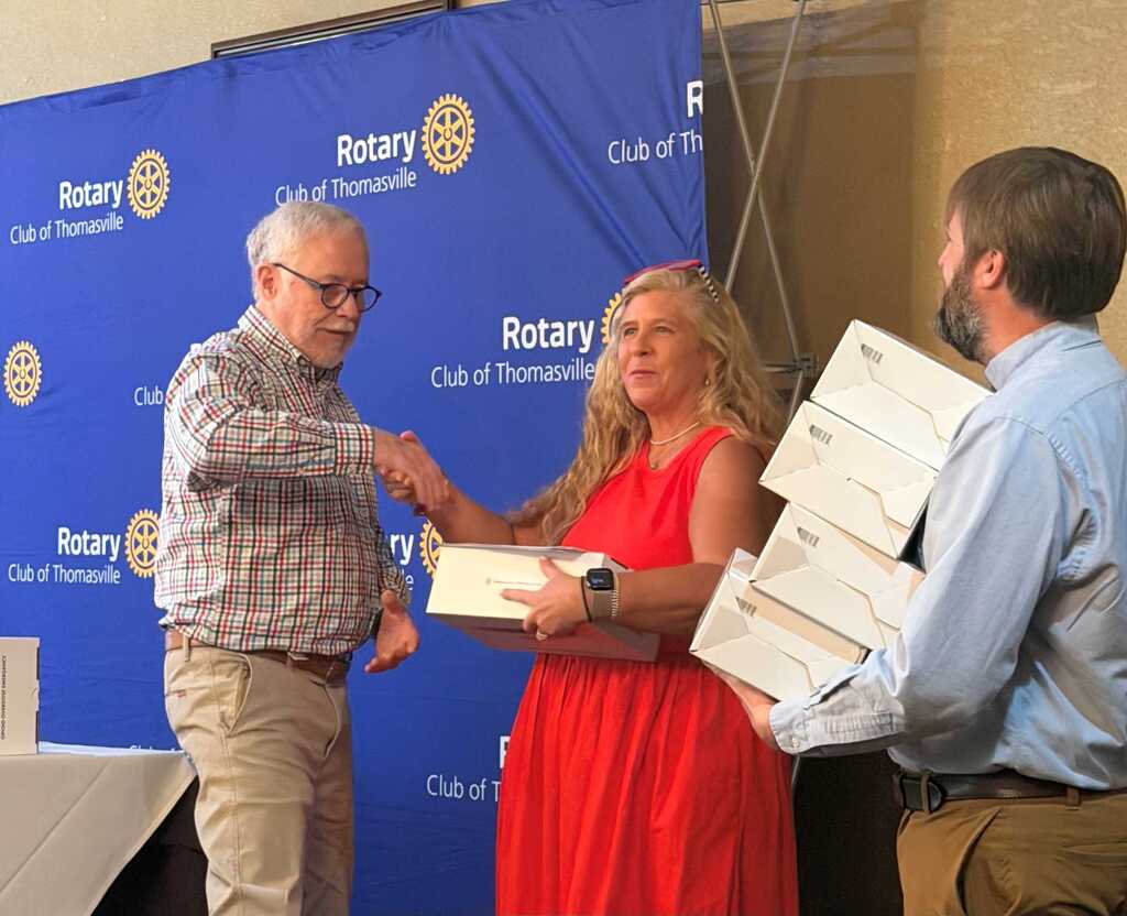 Three people stand in front of a blue banner. A man and a woman exchange documents.