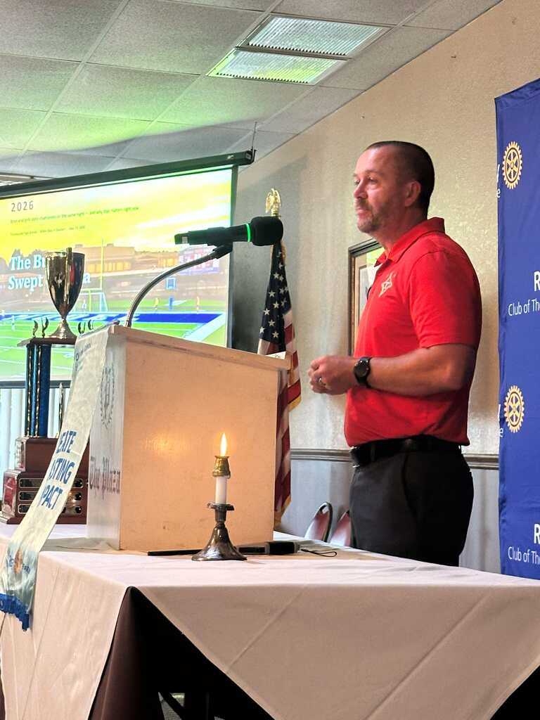 A man in red shirt stands at podium with a lit candle. A flag, microphone, and projector screen in background.