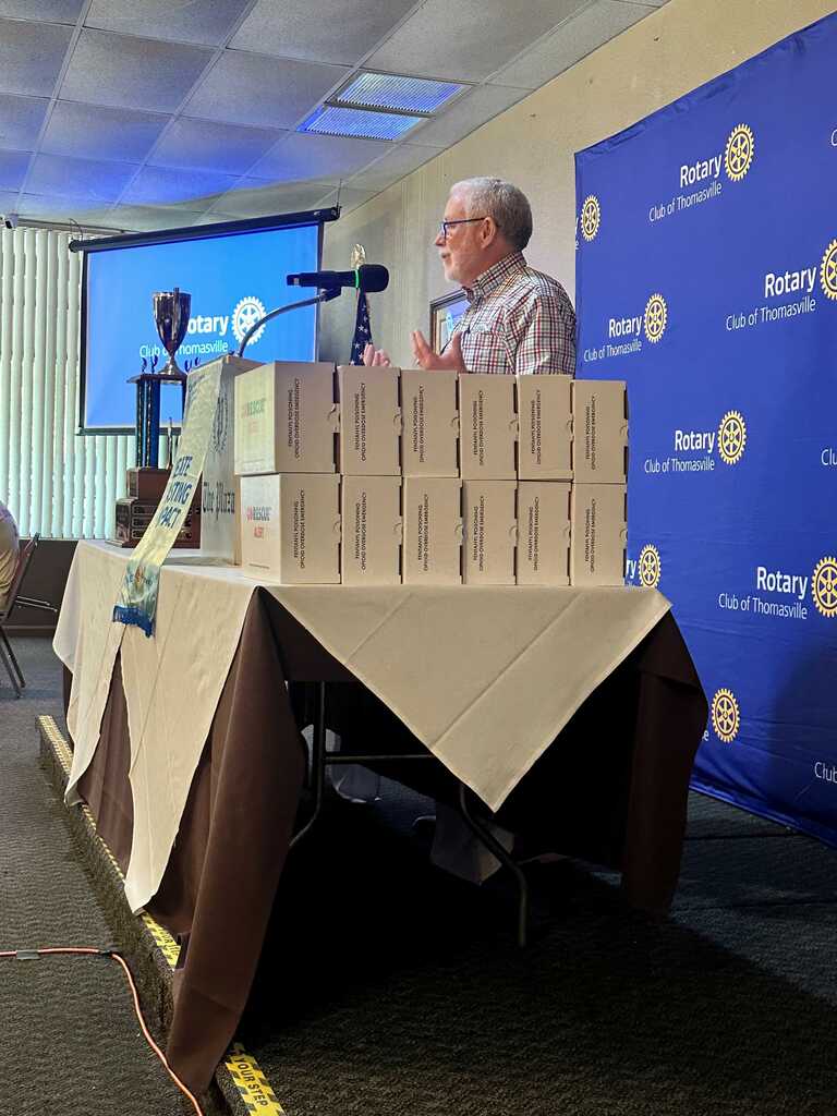 A man stands behind a table with boxes stacked on it, speaking into a microphone at an event.
