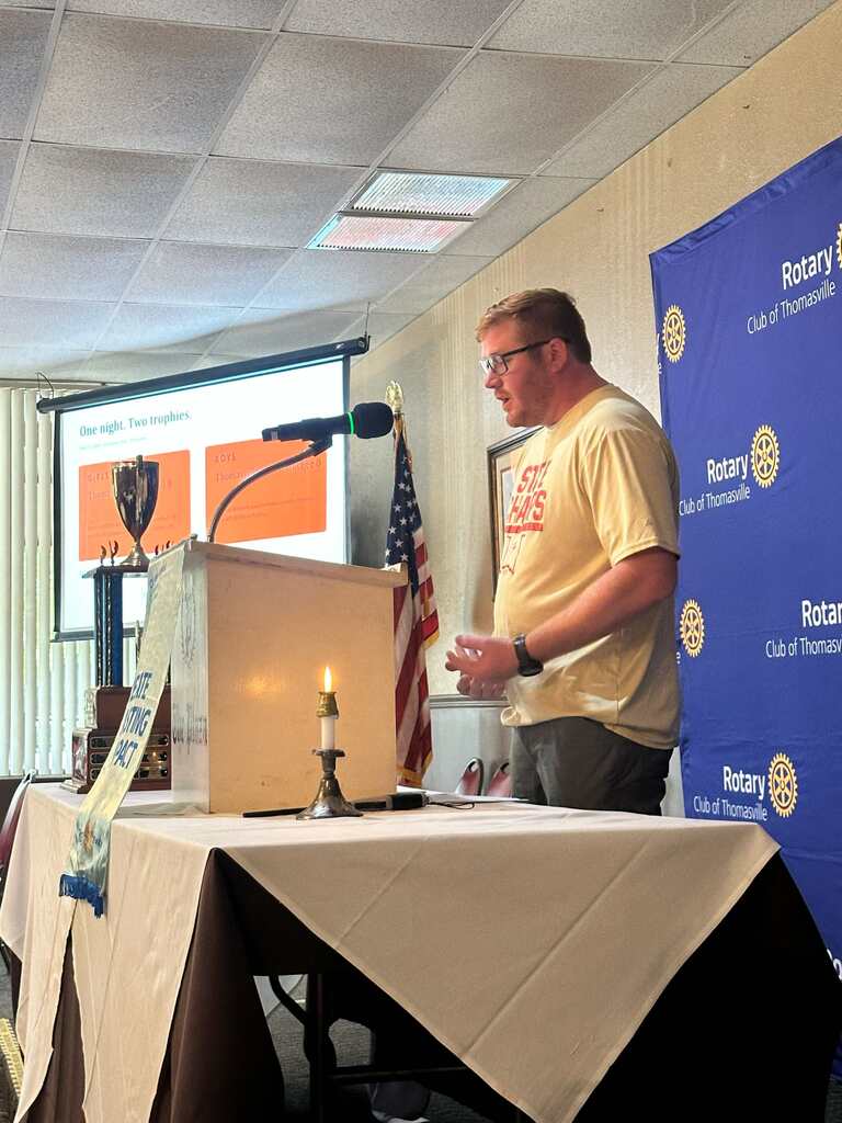 A man stands behind a podium with a candle and microphone, giving a presentation. A screen is behind him.