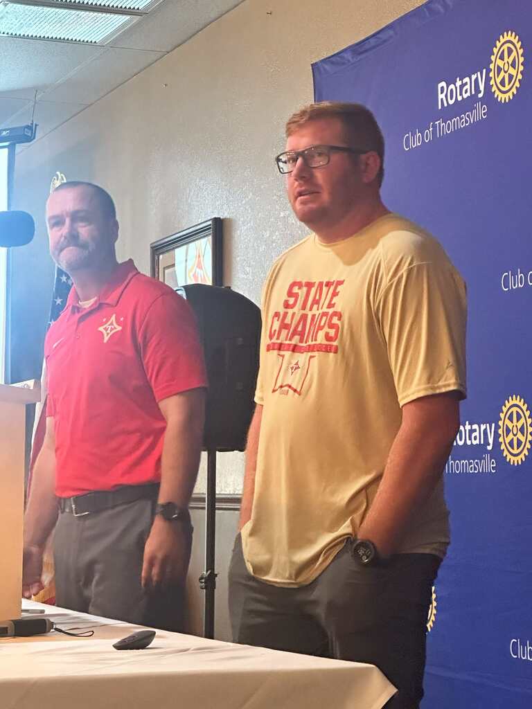 Two men, one in a red shirt and one in a yellow shirt, stand next to a table. Behind them, a wall displays a banner with the text "Rotary Club of Thomasville".