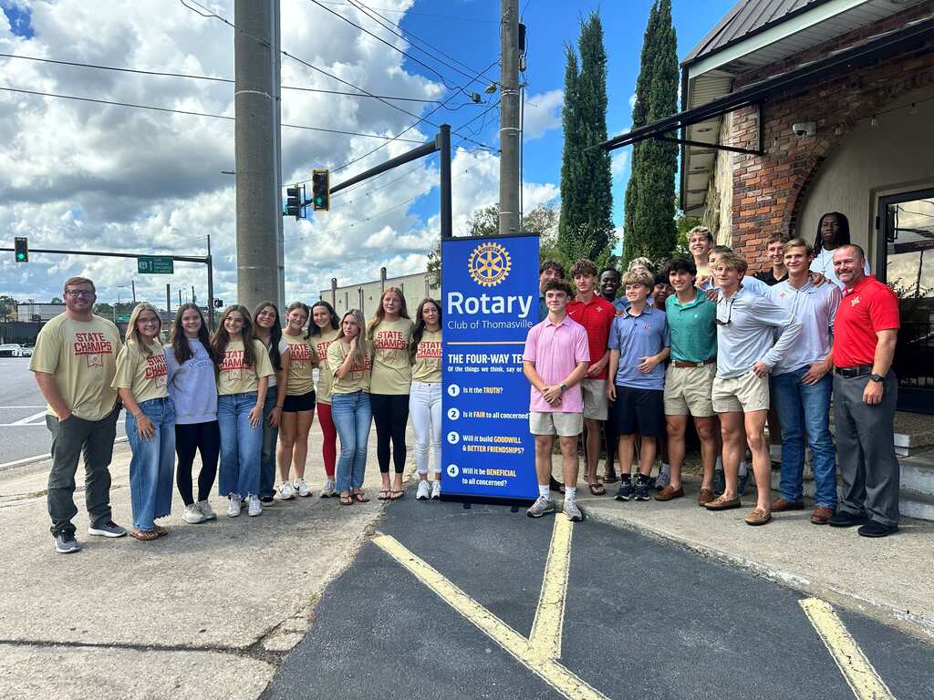Group of adults and teens standing outside, in front of a blue banner with the word "Rotary".