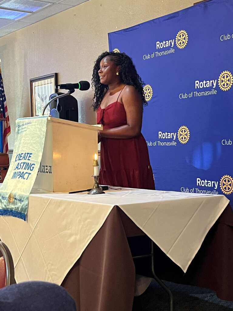 Woman in red dress stands at podium with microphone in front of Rotary Club banner. Table holds banner reading "CREATE LASTING IMPACT".