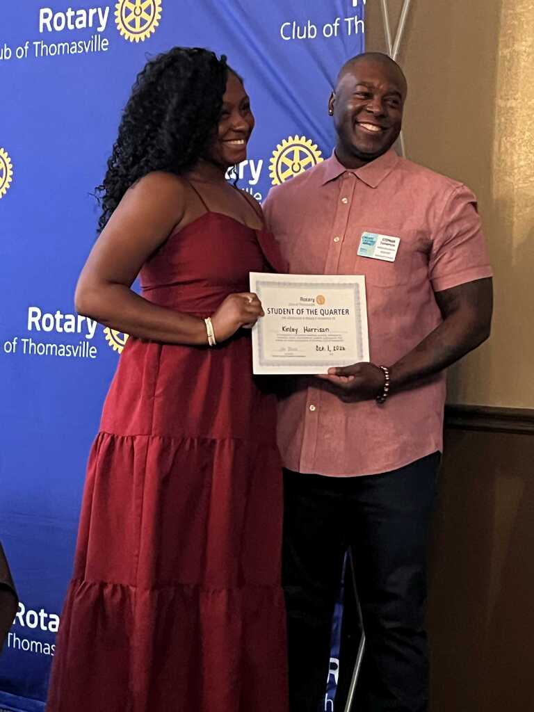 Person holding certificate stands next to another person with a smile on their face. Behind them is a blue banner.