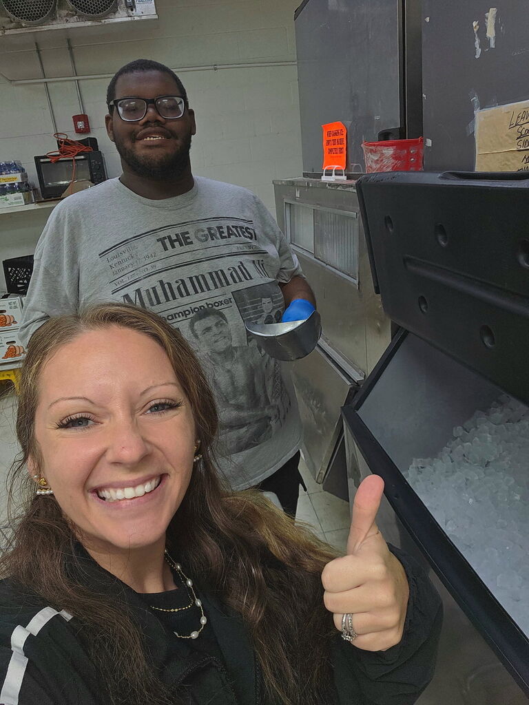 A smiling woman giving a thumbs up in front of an ice machine with a man wearing glasses in the background.
