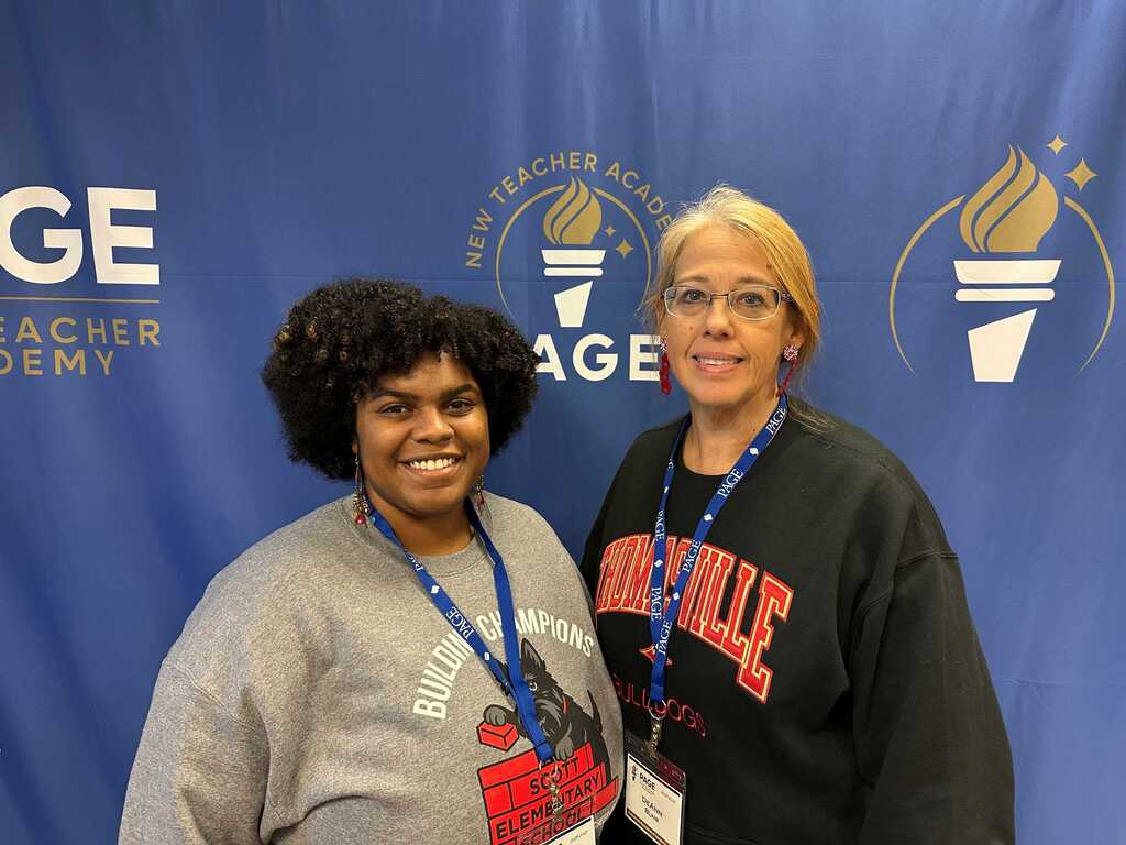 Two women stand side by side in front of a blue backdrop with logos. Both wear IDs on lanyards.