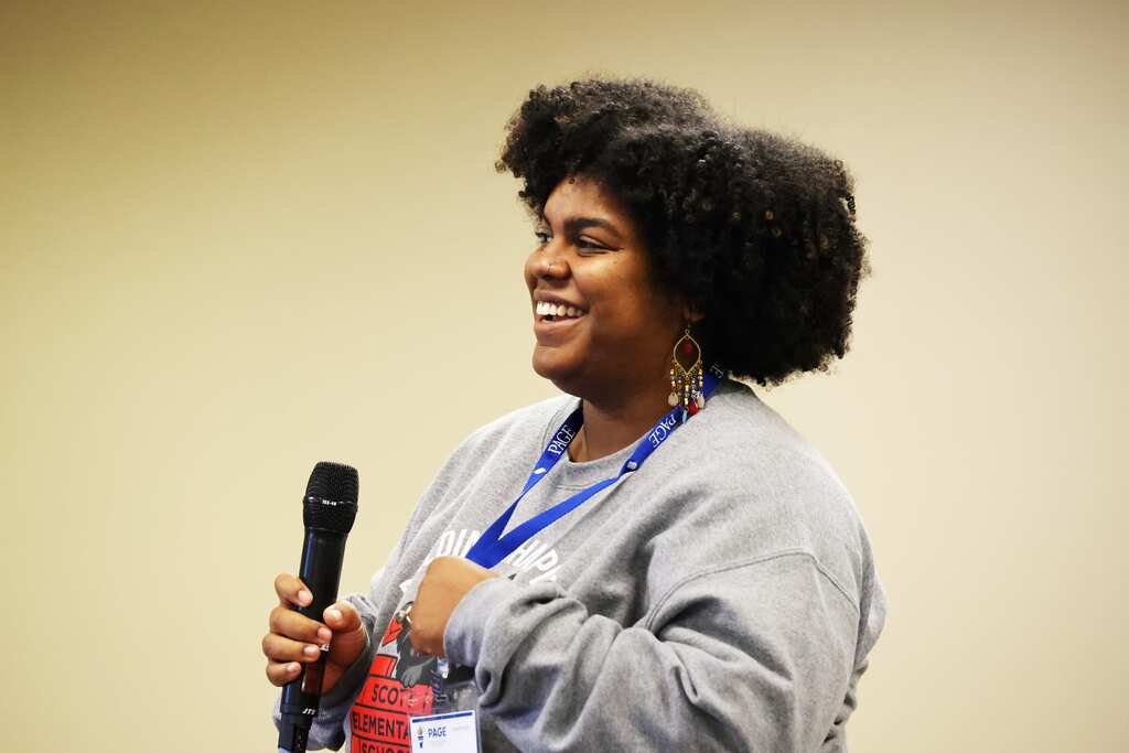 Woman with curly hair, wearing a gray shirt, holding a microphone with a blue lanyard around her neck.