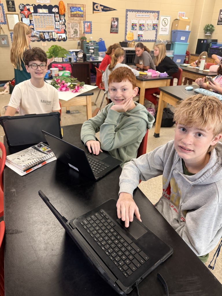 Three young boys sit at a table in a classroom with laptops in front of them.