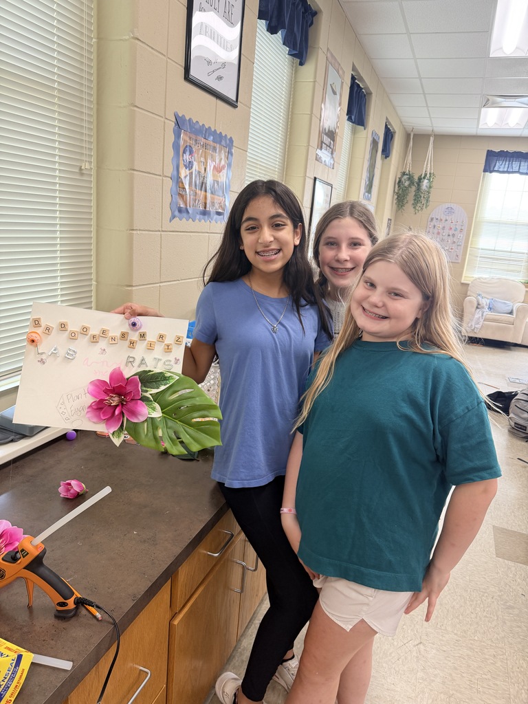 Three girls stand by a counter in a hallway, one holding a flower and a sign. Behind them, a wall has pictures and plants.
