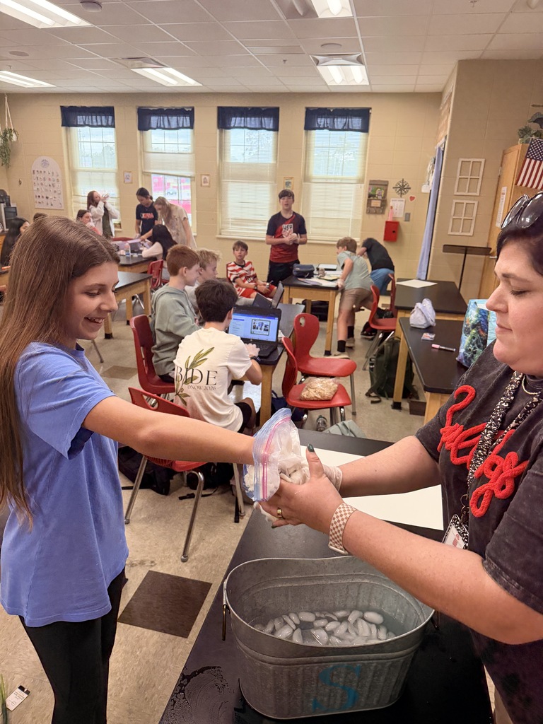 Two students are working together at a table in a classroom. One student is handing another student a bucket.