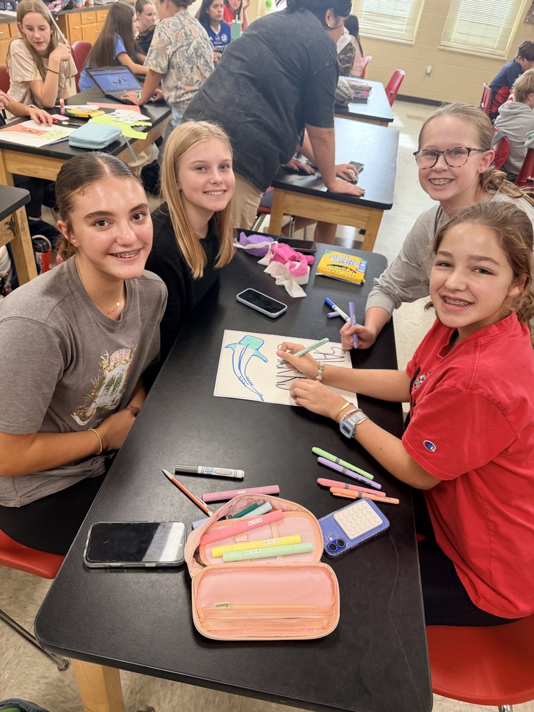 Students in a classroom seated at a desk, with colored pencils and a drawing. Some are smiling.
