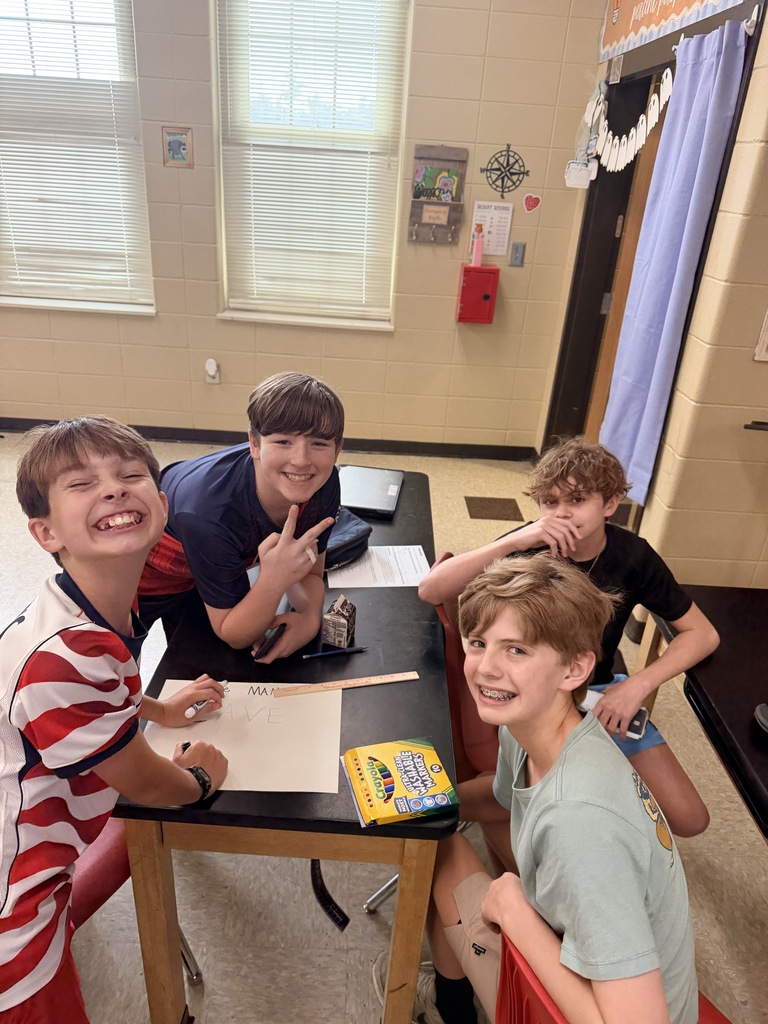 Four boys sit at a table, smiling. One holds a pencil. Papers and a box are on the table.