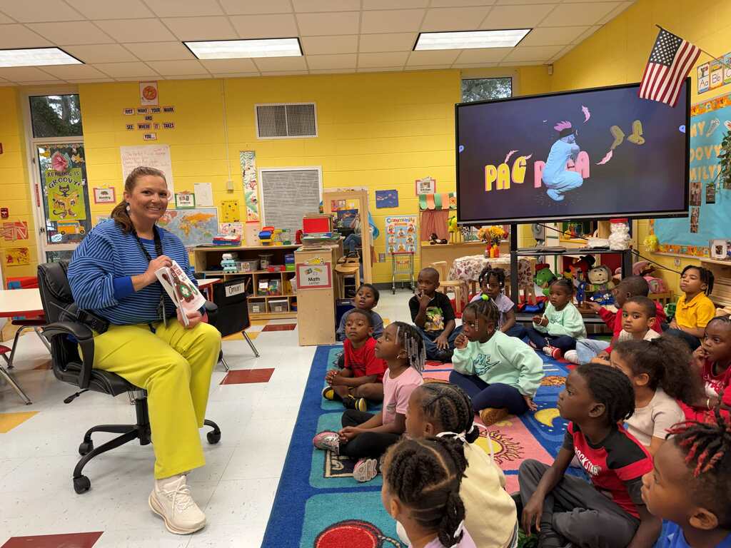 A woman reads a story to a classroom of children. They sit on a rug. Bright yellow walls and ceiling lighting.
