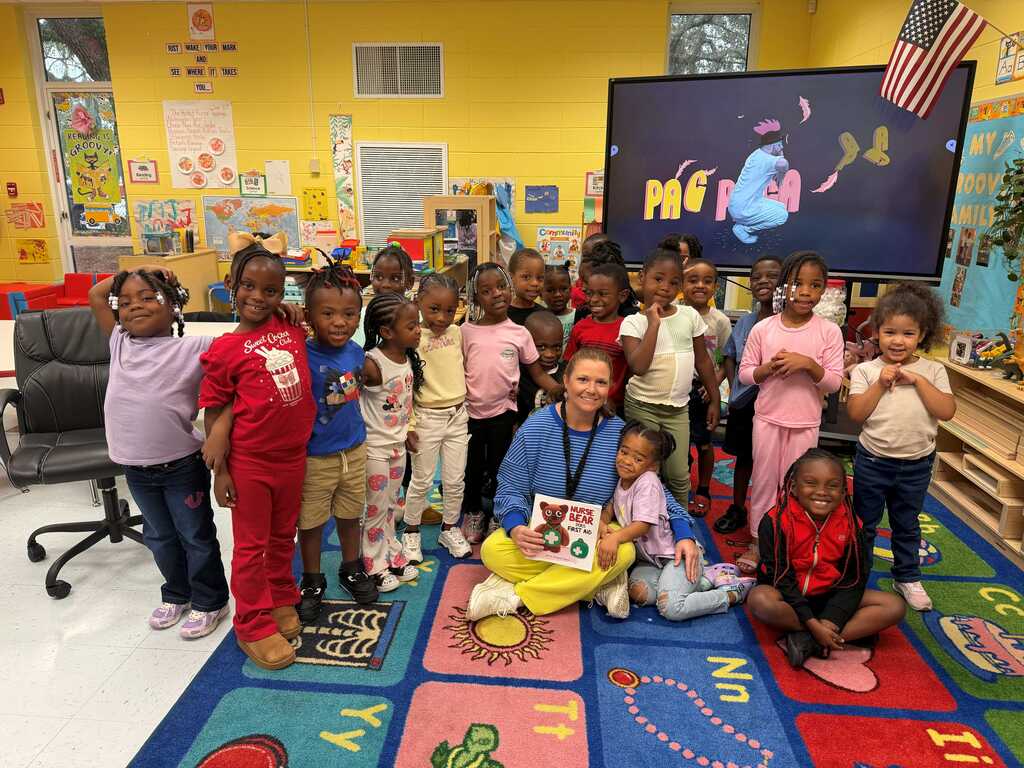 Group of young children in a classroom, some standing, some sitting on a colorful mat. A woman sits, holding a book.