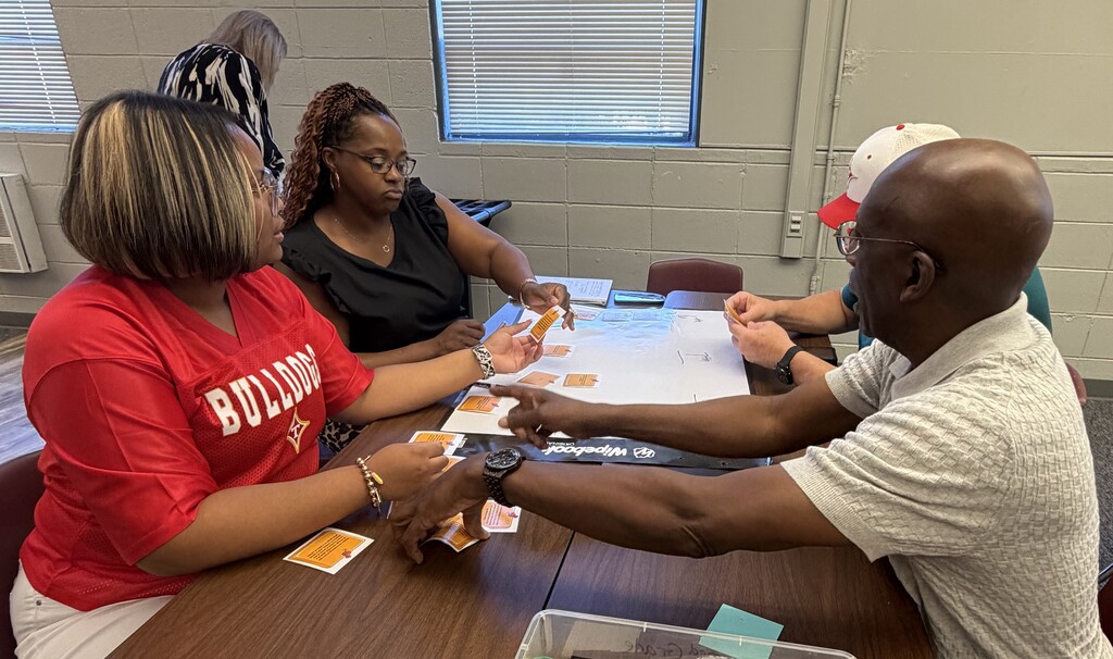 Three people seated at a table, exchanging game pieces. A woman wearing a red shirt with the word Bulldogs on it.