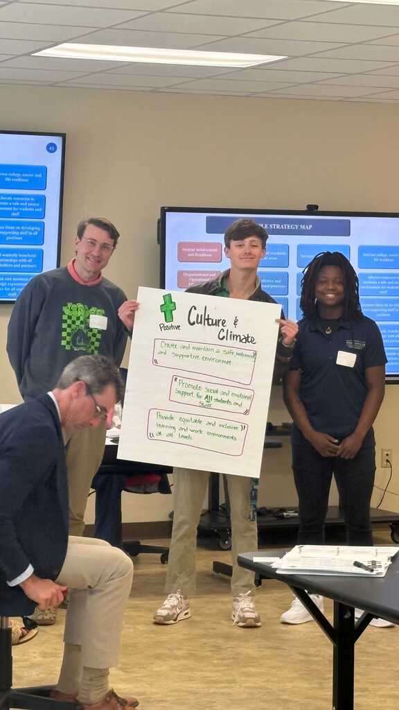 Four people standing in a room, one holding a large white board with text and images. Two screens on wall.