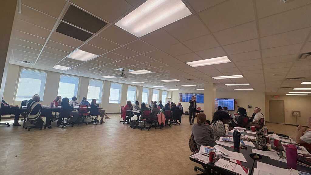 Group of people seated around tables in a room with windows and mounted ceiling lights.