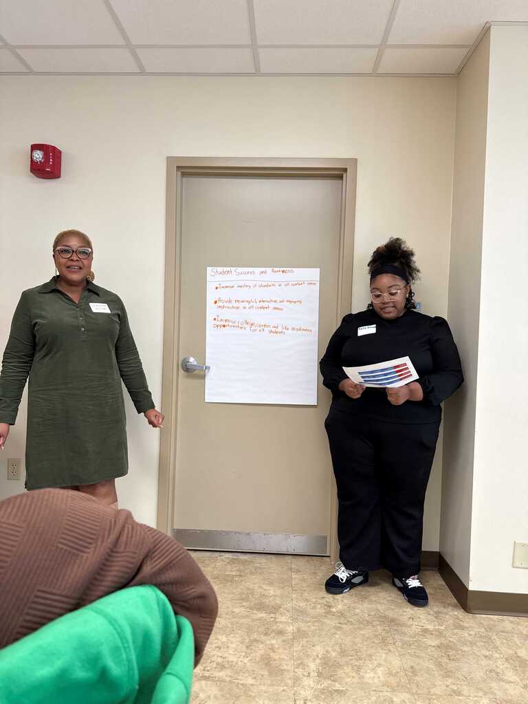 Two women stand in a hallway near a closed door, one holding a book, with a whiteboard on the door.